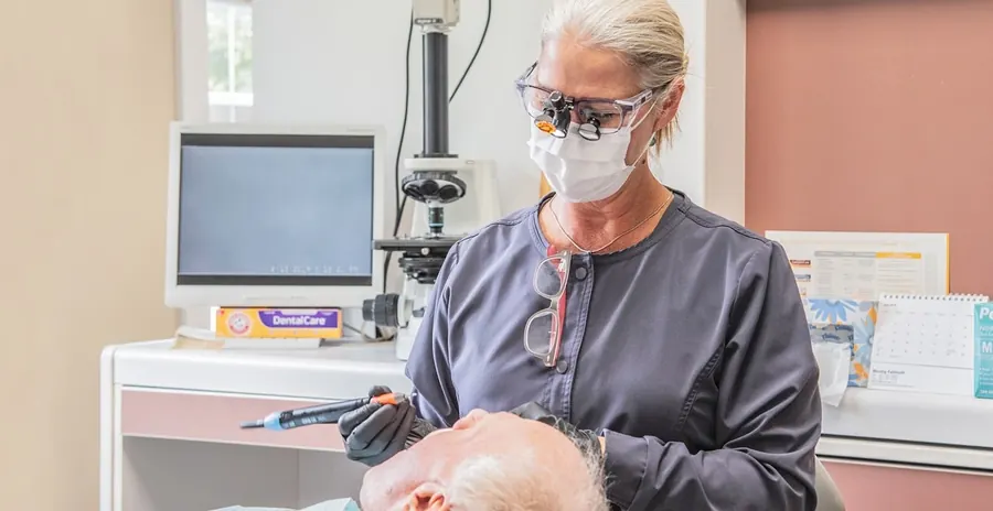 A Falmouth Dental Health hygienist in loupes cleaning a patient's teeth, with the monitor and microscope behind her.